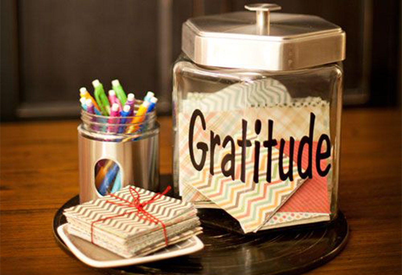 Um pote da gratidão em cima de uma mesa, com canetas coloridas e bloco de notas também coloridas ao lado. Foto: GateAwayToday