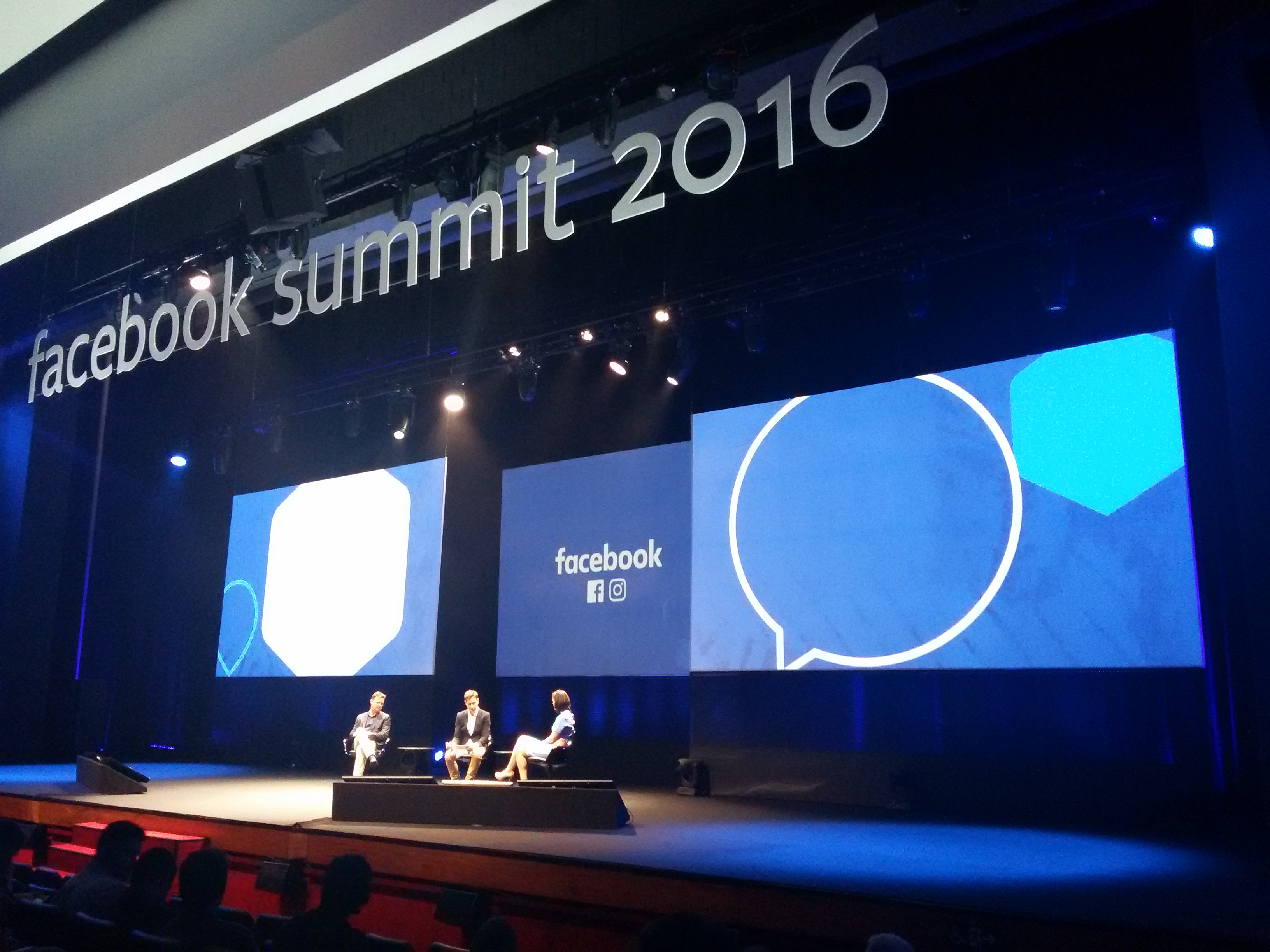 Três pessoas sentadas no palco do evento. Na parte superior do palco, há um letreiro com as palavras Facebook Summit 2016. O palco também tem três telões um ao lado do outro. 