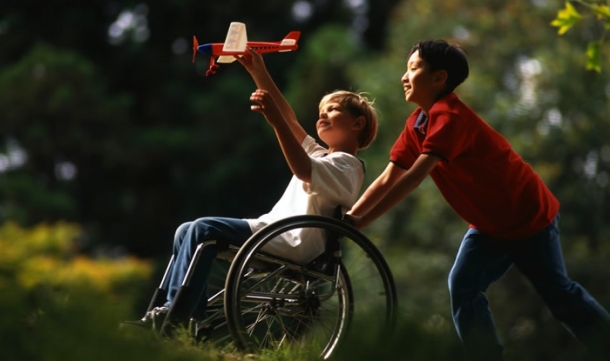 Menino empurra cadeira de rodas de um garoto com deficiência física, que brinca com um aviãozinho.