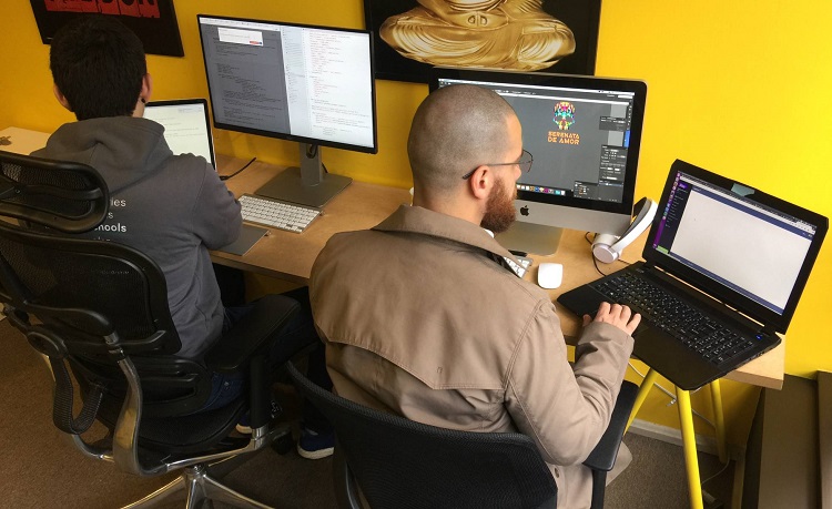 Foto de um escritório, com dois homens sentados lado a lado, trabalhando em computadores