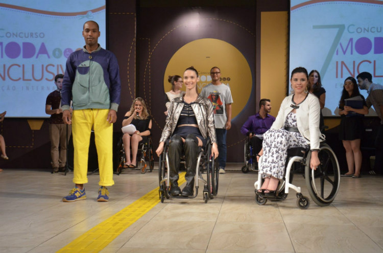 Foto de um desfile. Em uma passarela ampla, três modelos, um homem e duas mulheres cadeirantes desfilam suas roupas. Foto por Lincoln Yoshihashi