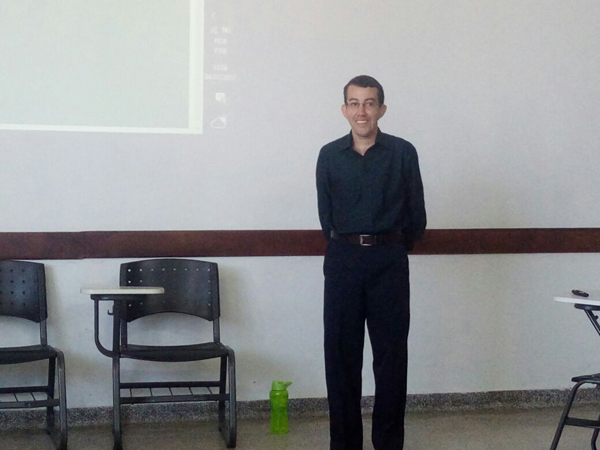Foto de um estudante, de aproximadamente 35 anos, em uma sala de aula. Ele está na frente de uma lousa digital, em pé, sorrindo e com as mãos para trás