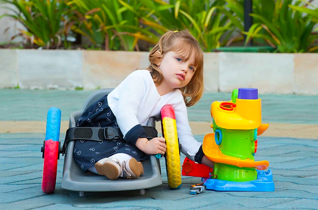 Foto de uma criança, de aproximadamente 2 anos, brincando. Ela está em uma cadeira de rodas adaptada, feita de um carrinho de plástico, quase na altura do chão, com duas rodas coloridas