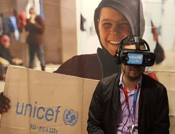 Foto de um jovem homem sentado, usando um fone de ouvido e óculos de realidade virtual. Atrás dele, há um banner da Unicef que mostra uma criança sorrindo