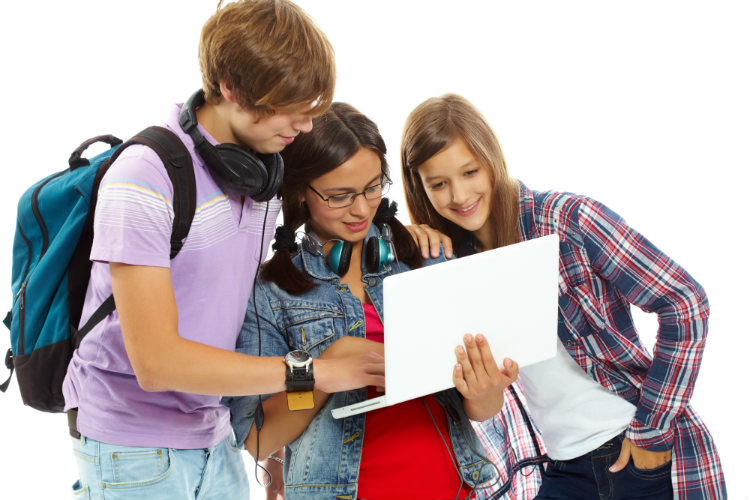 Foto de três adolescentes, um garoto e duas garotas, de aproximadamente 13 anos. Eles estão em pé e olham animados para a tela de um notebook, enquanto dois deles usam fones de ouvido pendurados no pescoço