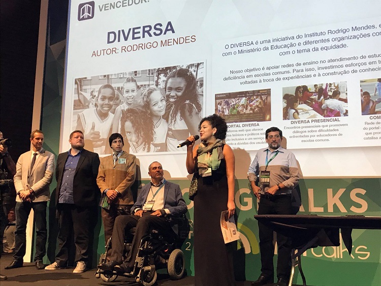 Aline Santos, à frente no palco com microfone, e cinco homens ao fundo, quatro em pé e um de cadeira de rodas.