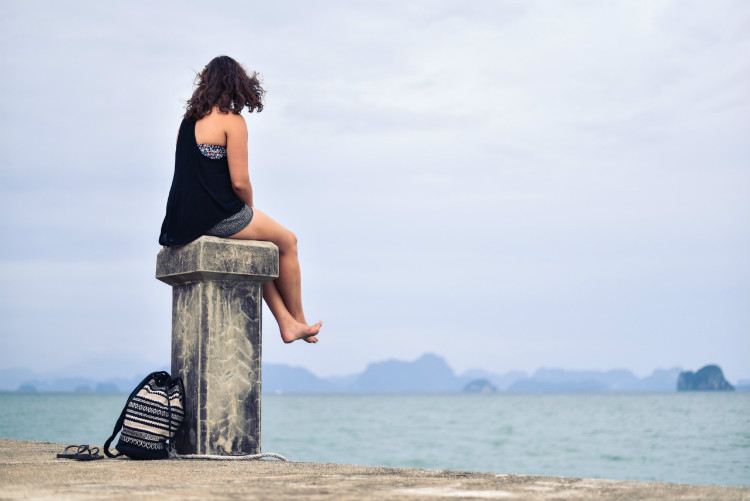 Foto de uma mulher jovem, de aproximadamente 20 anos, sentada em uma mureta à beira-mar. Ela está de costas para a imagem e observa o horizonte. Ao fundo, há um céu azul claro, ilhotas desfocadas e a superfície do mar