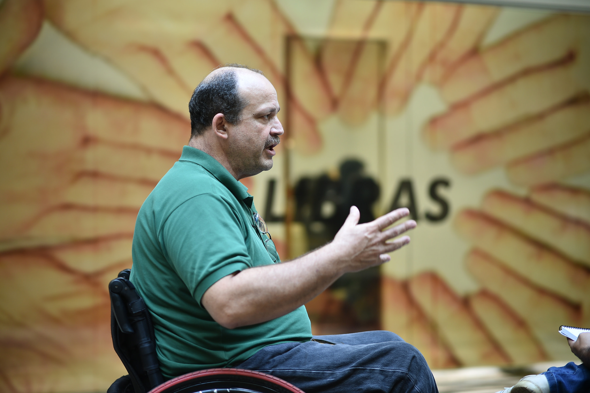 Homem sentado em cadeira de rodas. Ele usa uma camisa polo verde e gesticula enquanto fala. Ao fundo da imagem, em desfoque, há um banner com a palavra Libras.