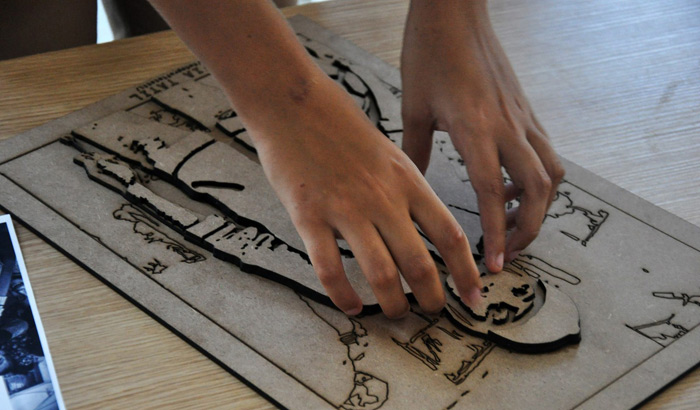 Foto de duas mãos tocando uma impressão em 3D. Ela é feita de madeira, tem sobreposições de camadas em alto relevo e reproduz a fotografia de uma pessoa