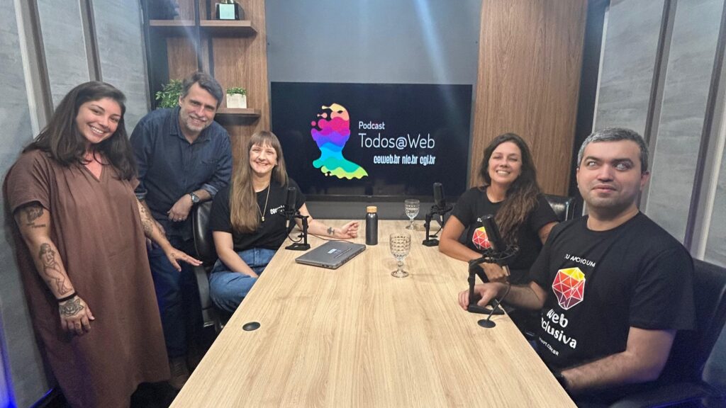 Foto de 5 pessoas ao redor de uma mesa retangular com microfones grandes, copos de água e notebook. Elas estão num ambiente fechado com paredes cinzas, móveis de madeira clara e, ao fundo, um monitor grande com o uma arte estilizada e o texto 