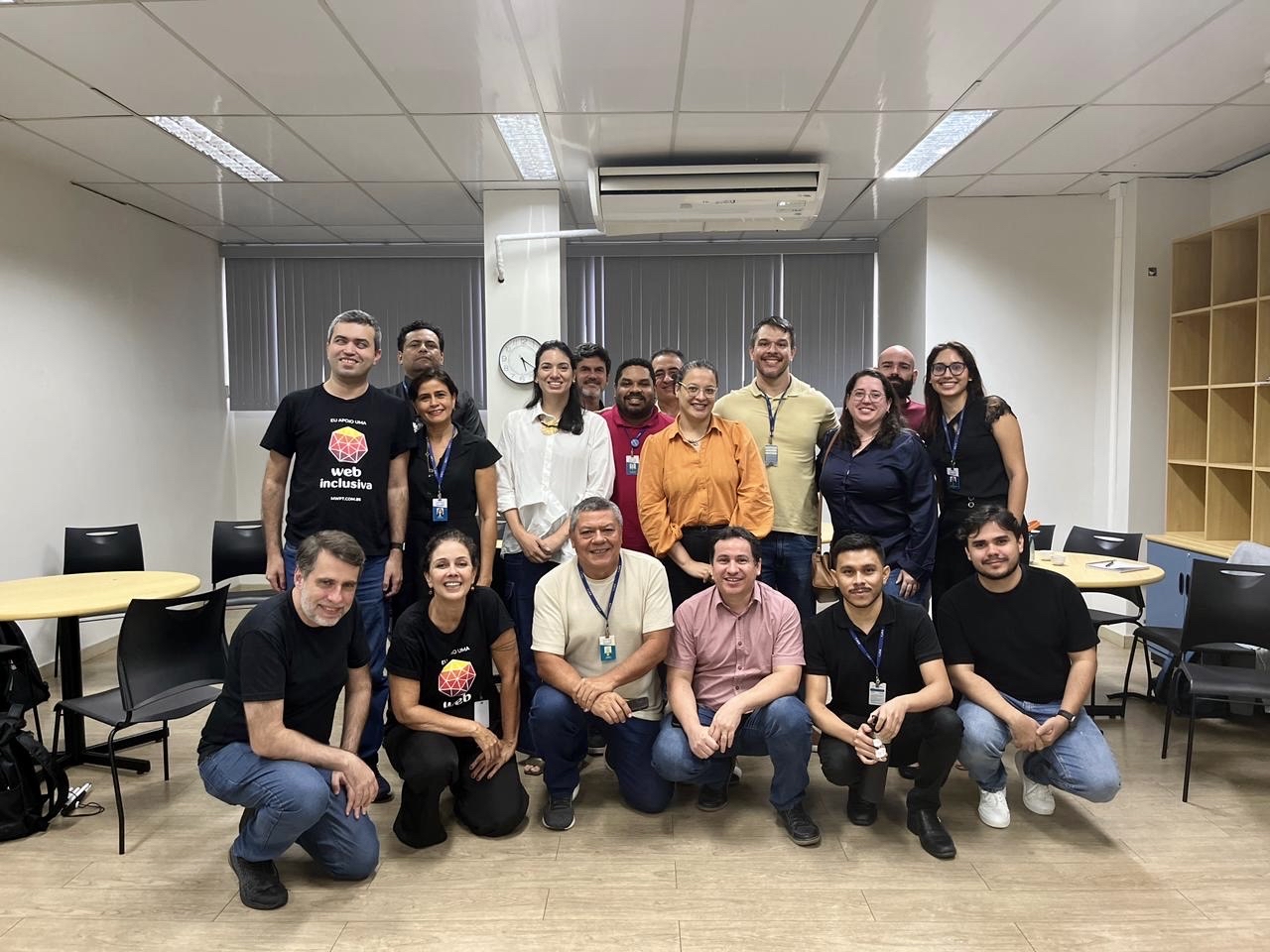 Foto de um grupo diverso de quase vinte pessoas, algumas em pé e outras abaixadas, em uma sala de aula.