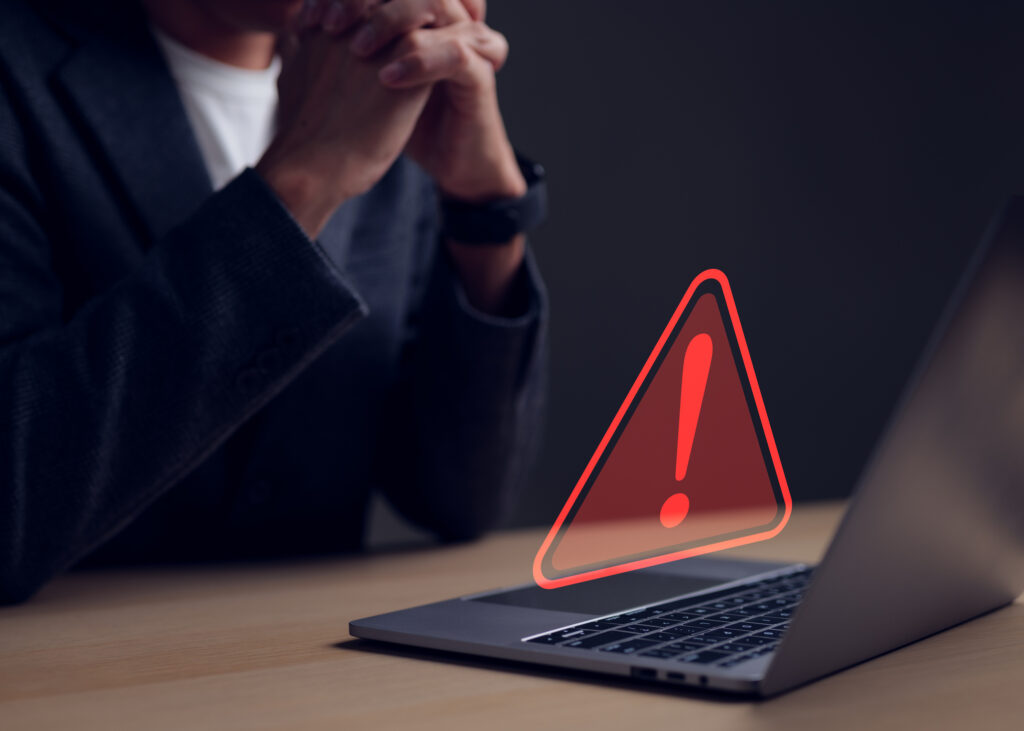 Homem de terno sentado à mesa diante de um notebook que exibe um sinal digital vermelho de alerta com ponto de exclamação.