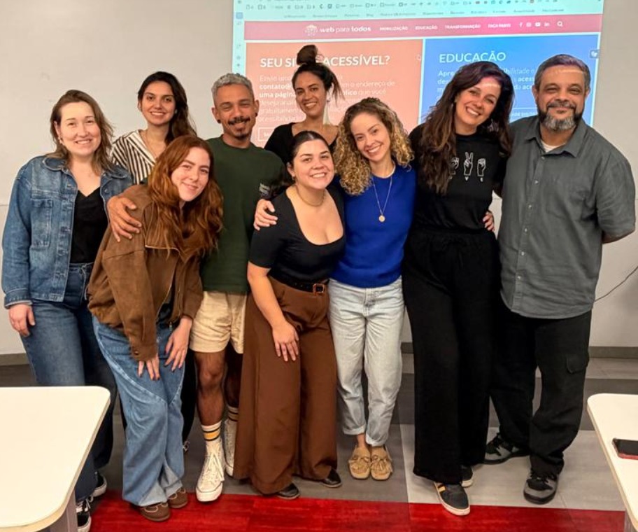 Foto de Simone Freire com JC Rodrigues e um grupo de estudantes em pé, lado a lado, abraçados e sorrindo. Ao fundo, projetado em uma parede branca, está a home do site do Movimento Web para Todos.