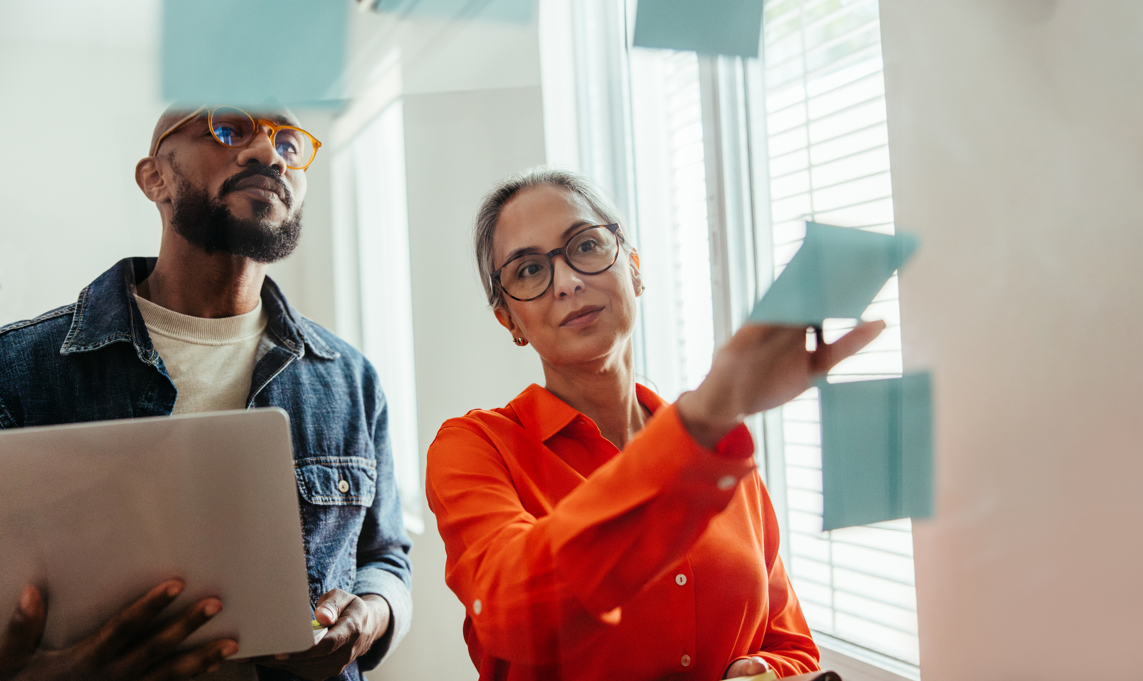 Homem e mulher em um escritório. Ela aponta para um vidro com notas adesivas coladas enquanto ele segura um laptop nas mãos.