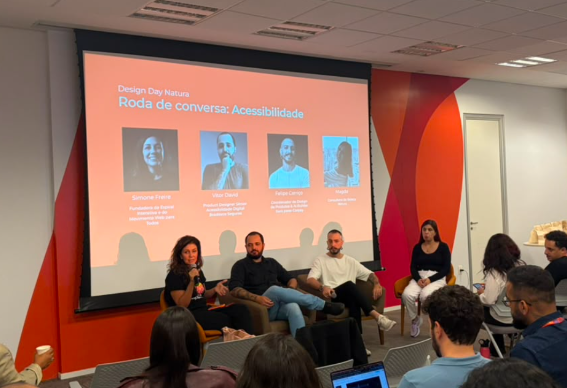 Foto de Simone Freire em painel realizado na Natura. Ela está sentada em um palco junto com outras três pessoas e na frente deles há uma plateia.