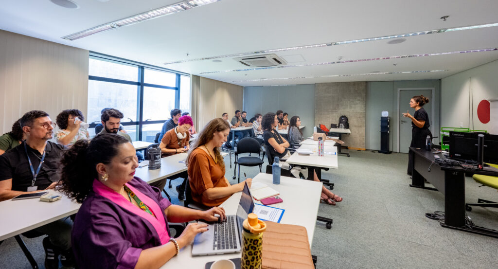 Foto de uma sala com um grupo diverso de pessoas sentadas em cadeiras. Na frente do grupo, em pé, está Simone Freire.