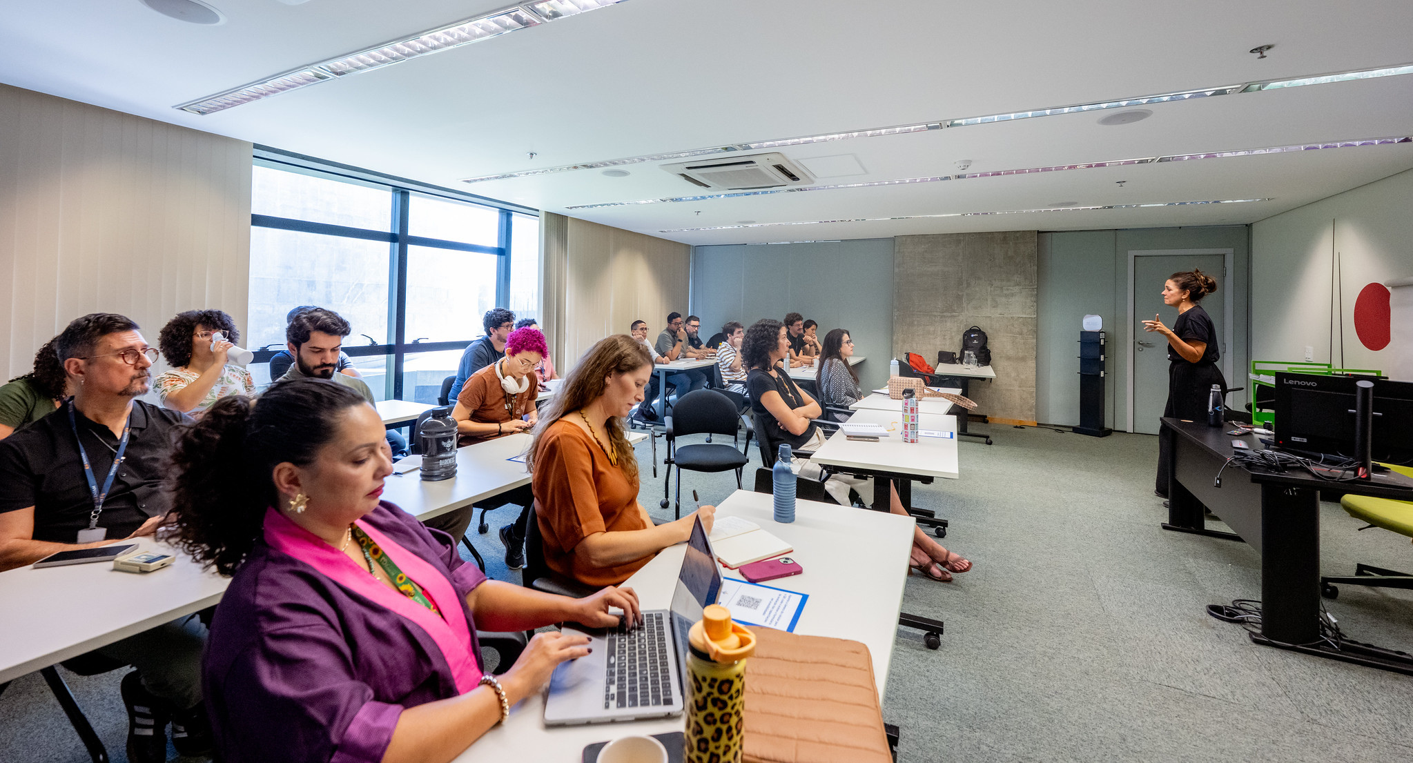 Foto de uma sala com um grupo diverso de pessoas sentadas em cadeiras. Na frente do grupo, em pé, está Simone Freire.